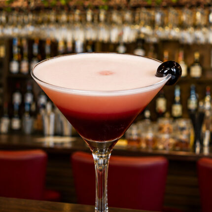 A signature layered pink and red cocktail with foam and a cherry garnish on the bar counter.