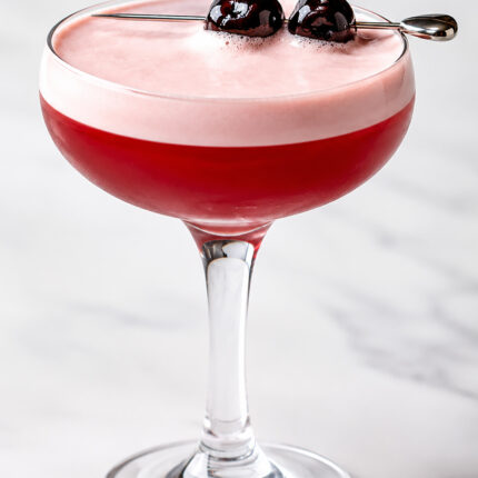An elegant red foam-topped cocktail garnished with skewered dark cherries in a coupe glass.