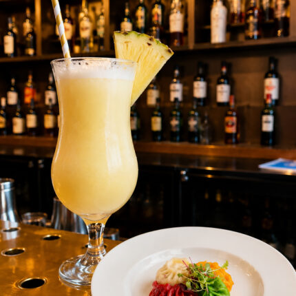 Tropical cocktail with a pineapple wedge and striped straw, placed next to a plate of vibrant red, green, and plain dumplings on a gold bar top.