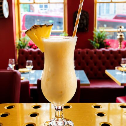 A frosty yellow blended cocktail garnished with a pineapple slice and a striped straw, sitting on a brass bar top with red booth seating.