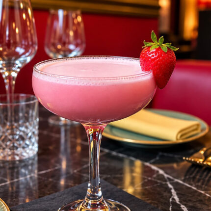 A creamy pink cocktail served in a glass coupe and garnished with a fresh strawberry, resting on a black marble dining table with wine glasses.
