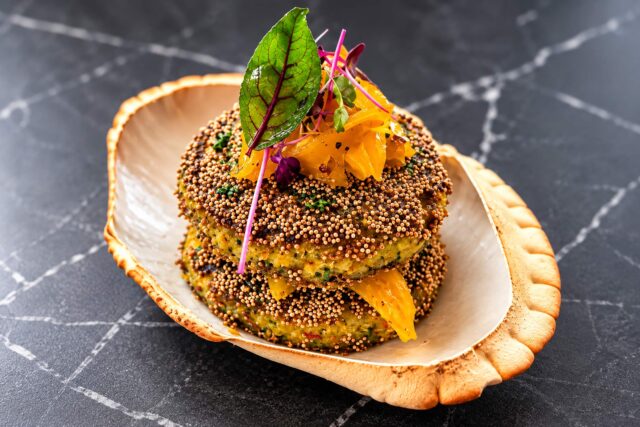 Two stacked, seed-crusted patties layered with yellow garnish and topped with microgreens, served in a unique shell-shaped ceramic bowl on a black marble table.