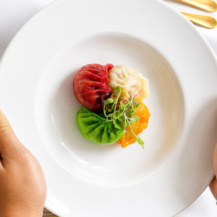 A diner's hands holding a white plate presenting four vibrantly coloured dumplings in red, white, green, and yellow, topped with microgreens.
