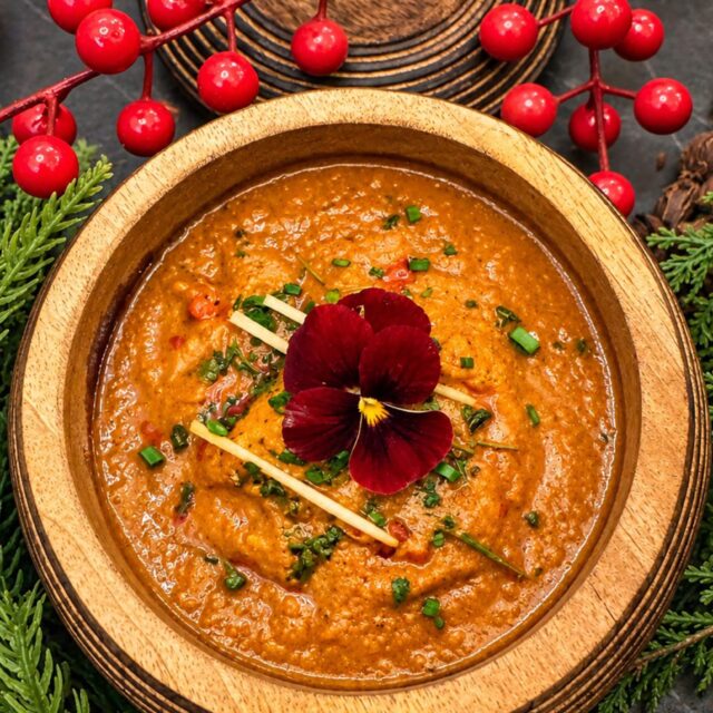 A rich Indian curry served in a carved wooden bowl, garnished with an edible red flower and surrounded by festive red berries and pine branches for the Christmas Special at Panas Restaurant.