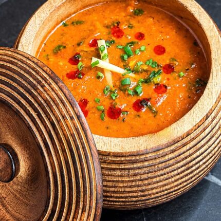 A vibrant orange curry or dal garnished with fresh green herbs, ginger matchsticks, and red chili oil droplets served in a carved wooden pot.
