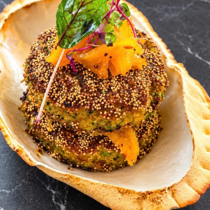 Two seed-crusted patties topped with pickled garnish and microgreens, creatively served inside a large decorative shell at Panas Restaurant.