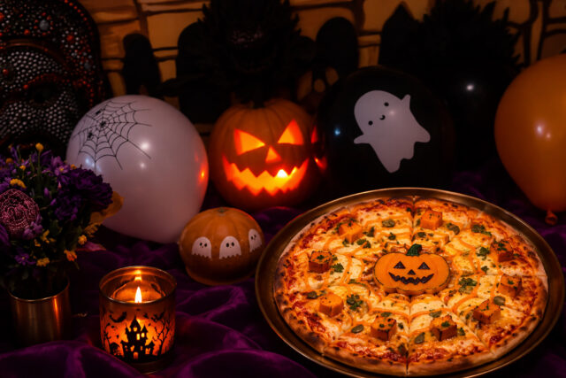 A festive Halloween-themed pizza decorated with a central jack-o'-lantern face and pumpkin cubes, surrounded by spooky balloons, lit pumpkins, and candles at Panas Restaurant.