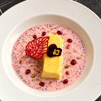 A yellow dessert resting in a pink milky base with sago pearls, garnished with pomegranate seeds, pistachios, and a decorative red leaf.