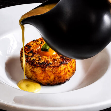 A dark jug pouring a rich yellow sauce over a golden, pan-seared round patty garnished with a single green leaf at Panas Restaurant.