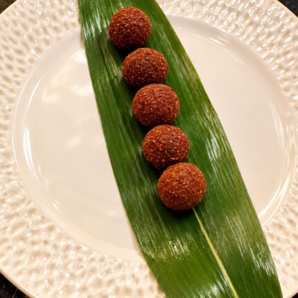 Five perfectly round, spiced Indian croquettes lined up elegantly on a long green leaf over a textured white plate at Panas Restaurant.