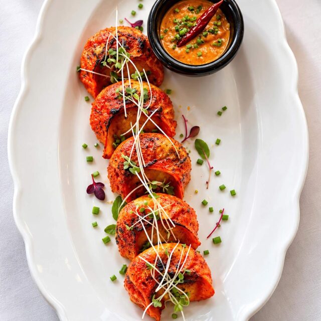 Five vibrant tandoori momo dumplings garnished with microgreens, served with a spicy chilli dipping sauce on a white platter at Panas Restaurant.